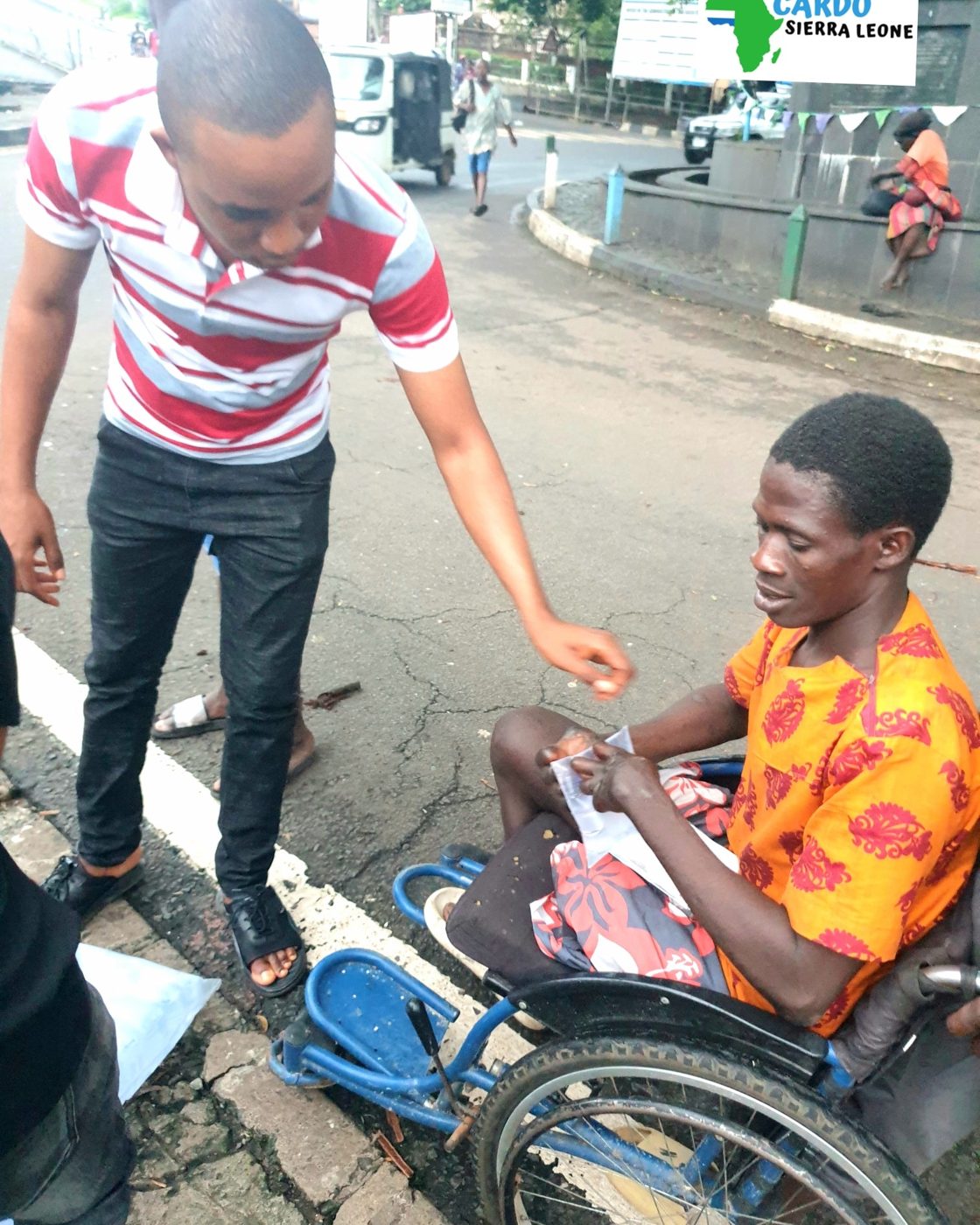 A CARDO volunteer supporting a man in a wheelchair during an outreach in Freetown